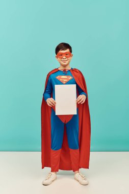 full length of preteen asian boy in superhero costume with mask and cloak standing with blank paper and looking at camera on blue background, World child protection day concept  clipart