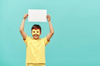 smiling multiracial boy in yellow superhero costume with mask holding blank paper above head on blue background, International children's day concept  clipart
