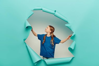 cheerful redhead girl in t-shirt smiling and looking at blue torn paper hole on white background, International Child Protection Day concept  clipart