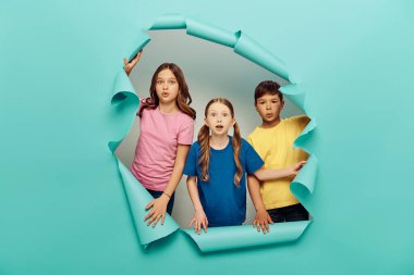 Shocked multiethnic preteen kids in colorful t-shirts looking at camera while celebrating child protection day behind hole in blue paper background clipart