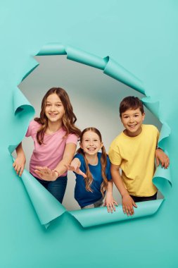 Renkli tişörtler giymiş pozitif çoklu etnik çocuklar kameraya doğru uzanırken uluslararası çocukların mavi kağıt arka plandaki deliğe yakın günlerini kutluyorlar.