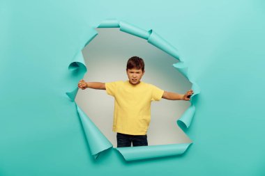 Angry multiracial boy in yellow t-shirt looking at camera during international child protection day while standing behind hole in blue paper on white background clipart