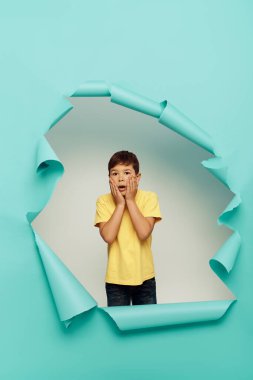 Shocked multiracial boy in yellow t-shirt touching cheeks and looking at camera during world child protection day while standing behind hole in blue paper background clipart