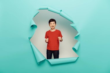 Cheerful asian preteen boy in red t-shirt dancing and looking at camera during child protection day celebration behind hole in blue paper white background clipart