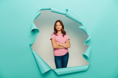 Smiling preteen girl in casual clothes crossing arms and looking at camera during child protection day celebration behind hole in blue paper background clipart