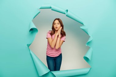Shocked preteen girl in pink t-shirt looking at camera during international children day celebration while standing behind hole in blue paper background clipart
