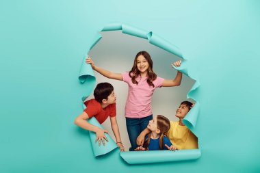 Smiling interracial kids in colorful clothes looking at friend while celebrating international children day behind hole in blue paper background clipart