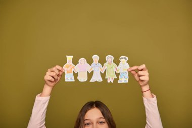 Cropped view of preteen girl holding hand-drawn paper characters above head and looking at camera during child protection day celebration on khaki background clipart