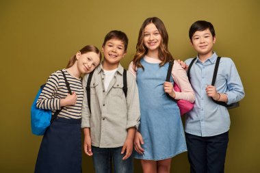 Positive interracial and preteen children in casual clothes holding backpacks and looking at camera during child protection day celebration on khaki background clipart