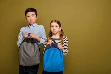 Scared multiethnic preteen kids in casual clothes holding backpacks and looking at camera together during international child protection day celebration on khaki background clipart