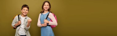 Carefree multiracial boy with backpack showing yes gesture at camera near friend during child protection day celebration on khaki background with copy space, banner  clipart