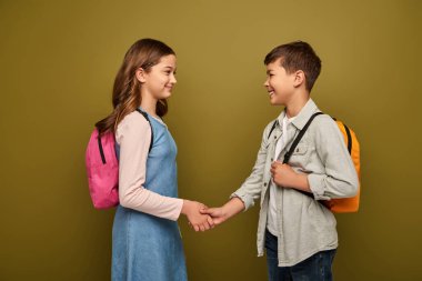 Smiling multiethnic schoolkids with backpacks shaking hands and looking at each other during international child protection day celebration on khaki background clipart