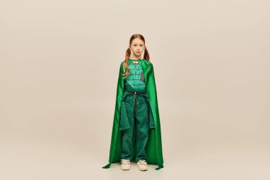 Küresel Çocuk Koruma Günü 'nde gri arka planda dikilirken yeşil süper kahraman kostümlü ve pelerinli kızıl saçlı kız kameraya bakıyor. 