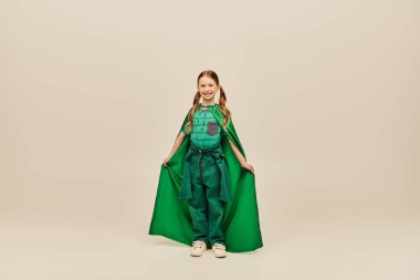 Cheerful redhead kid in green superhero costume holding cape and looking at camera while standing on grey background in studio during international children`s day celebration clipart