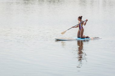 slender african american woman in colorful swimsuit holding paddle while standing on knees and sailing on sup board on river during weekend on summer day clipart