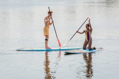 cheerful african american woman in colorful swimsuit kneeling on sup board near redhead man sailing with paddle while spending time on river in summer clipart