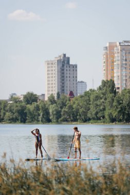 Mayo giyen sporcu bir çift ellerinde raketlerle yelken açarken gölde manzaralı şehir manzarası ve bulanık ön planda bitkilerle vakit geçiriyorlar.