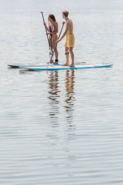 full length of young redhead man in swim shorts and brunette african american woman in colorful swimsuit sailing on sup boards on calm river on summer day clipart