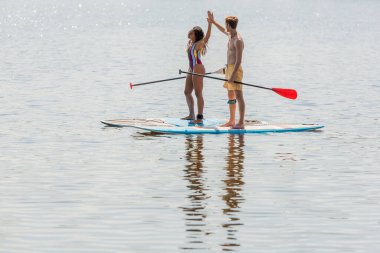 positive and sportive redhead man and african american woman in colorful swimwear holding paddles and giving high five while sailing on sup boards on lake in summer clipart