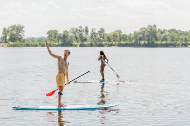 cheerful redhead man looking away and pointing with hand near sportive african american woman in colorful swimsuit sailing on sup board on summer day with picturesque riverside on background clipart