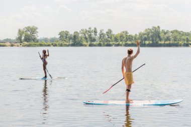 active interracial couple in colorful swimwear looking at each other and waving hands while sailing on sup boards on picturesque lake with green shore clipart
