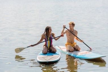carefree african american woman and young, cheerful redhead man in colorful swimwear spending summer vacation on lake and sailing on sup boards with paddles clipart