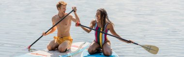 young and happy redhead man and excited african american woman in striped swimsuit kneeling on sup boards while sailing on lake during water recreation in summer, banner clipart