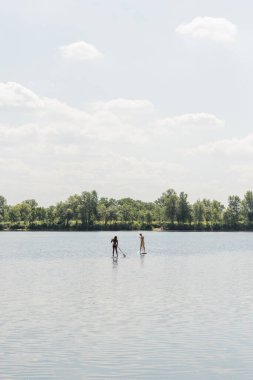 view from afar of sportive interracial couple sailing on sup boards while spending weekend on lake with green picturesque shore under cloudy sky, outdoor activity, summer vibes clipart