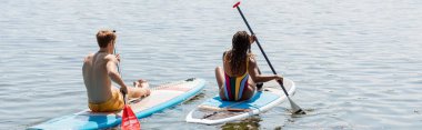 back view of young redhead man and sportive african american woman in striped swimsuit sitting on sup boards and sailing on lake on summer day, banner clipart