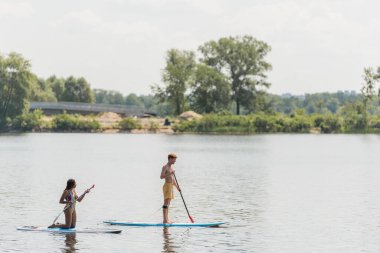 Genç sporcu adam ve Afro-Amerikan kadının dizlerinin üzerinde duran ve yelkenli yelkenliyle yeşil bankayla nehirde yüzen