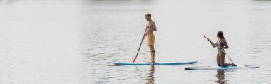 full length of redhead and sportive man sailing on sup board near african american woman in striped swimsuit on calm river during summer weekend, banner clipart