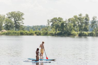 active multiethnic couple in colorful swimwear sailing on sup boards with paddles while spending summer vacation day on river with green trees on scenic bank clipart