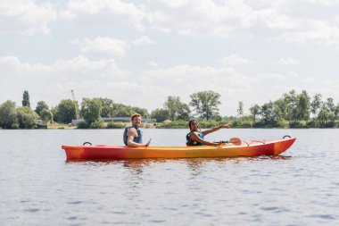carefree african american woman in life vest pointing ahead and smiling at camera while sailing in sportive kayak with redhead friend during summer weekend on river clipart