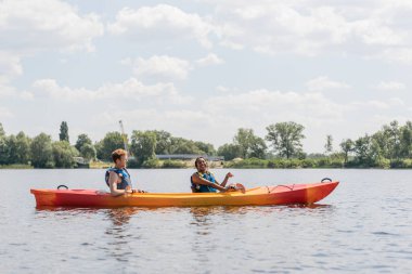 Neşeli Afro-Amerikalı kadın ve kızıl saçlı genç adamın yan görünüşü nehirde sportif bir kanoyla yelek giyiyor mavi ve bulutlu gökyüzünün altında yeşil kıyısı var.