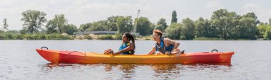 side view of sportive and interracial couple in life vests sailing in kayak during summer water recreation on river with green picturesque bank, banner clipart