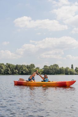 carefree interracial couple in life vests giving high five while spending summer weekend on river and sailing in sportive kayak along green riverside under blue sky with white clouds clipart