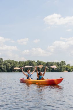 couple of cheerful interracial friends in life vests holding paddles in raised hands while sitting in kayak on picturesque lake under blue cloudy sky with green riverside on background clipart