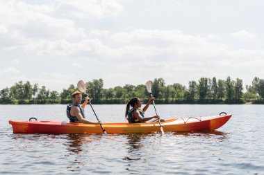 side view of smiling redhead man and brunette african american woman in life vests spending summer weekend by sailing in kayak on river under cloudy sky in summer clipart