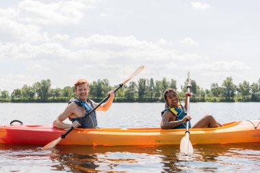 pretty african american woman and young redhead man in life vests smiling at camera while spending summer weekend on river and sailing in kayak with paddles under blue cloudy sky clipart