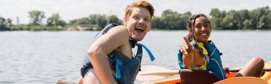 young and excited redhead man with open mouth looking at camera near overjoyed african american woman in life vest pointing with finger while sailing in kayak on summer day, banner clipart