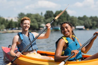 pleased african american woman and cheerful redhead man in life vests holding paddles while sailing in sportive kayak during summer weekend on lake on blurred background clipart
