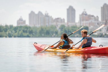 redhead man and african american woman in life vests spending time on river by sailing in sportive kayak and enjoying picturesque cityscape on blurred background clipart