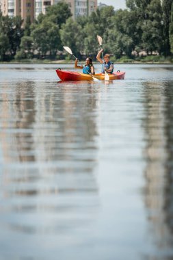 young active man and african american woman in life vests sailing in sportive kayak with paddles on blurred water surface with green riverside on background clipart