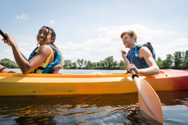 smiling and pretty african american woman sailing in sportive kayak with active redhead man in life vest during summer vacation on river with green bank clipart