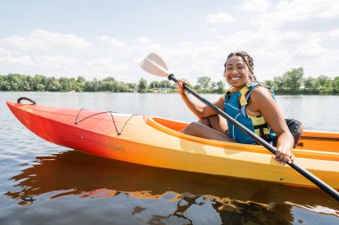 Çok neşeli Afro-Amerikan kadını sportif bir kanoyla kürek çekiyor ve gölün arka planında yeşil resimli sahile gülümsüyor.
