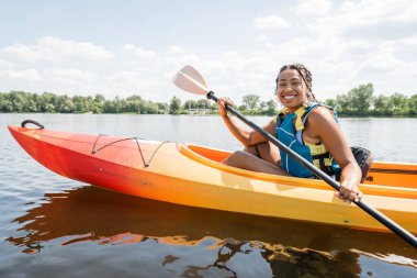 overjoyed african american woman in life vest paddling in sportive kayak and smiling at camera on lake with green picturesque shore on background in summer clipart