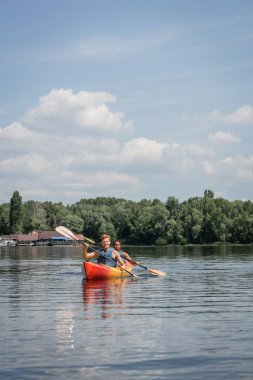Spor hayatındaki çok ırklı çift yaz tatilinde mavi bulutlu gökyüzünün altında yeşil renkli bir bankayla nehirde kano üzerinde kürek çekiyorlar.