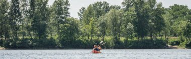 view from afar of interracial active couple in life vests sailing in sportive kayak with paddles near riverside with green trees on summer day, banner clipart