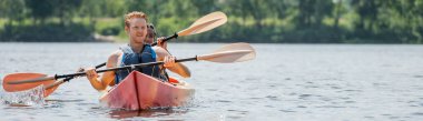 young and sportive redhead man in life vest looking away while holding paddle and sailing on kayak with african american woman on river in summer, banner clipart