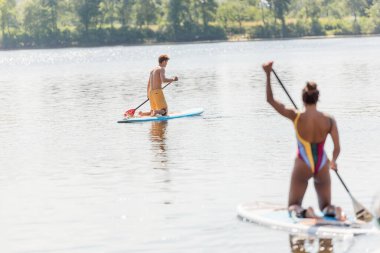 Genç sporcu adam, Afrika kökenli Amerikalı bir kadının yanındaki pitoresk gölde yelken açıyor. Renkli mayo giymiş, yazın bulanık ön planda kürek çekiyor.